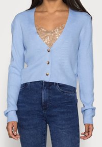 Light blue ribbed cardigan with a deep V-neck, five wooden buttons, and long sleeves. Worn over a lace top and paired with dark jeans.