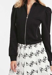 Chaqueta negra corta con cremallera frontal y mangas abullonadas, combinada con una falda blanca plisada que presenta un patrón de logo negro repetido.