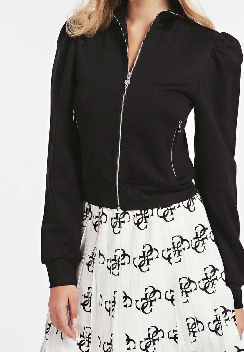 Chaqueta negra corta con cremallera frontal y mangas abullonadas, combinada con una falda blanca plisada que presenta un patrón de logo negro repetido.