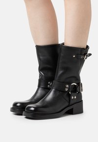 Botas de cuero negras hasta el tobillo con punta redondeada, cinta ancha, acentos de herrajes plateados y un tacón bajo en bloque. Textura suave con un ligero brillo.