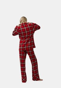 Persoon met lang blond haar die de kraag aanpast, gekleed in een rood geruit jasje en bijpassende broek, blootsvoets, staand tegen een effen achtergrond.