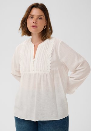 Femme aux cheveux bruns mi-longs, portant des boucles d’oreilles créoles dorées et un chemisier blanc à manches longues avec des détails en dentelle sur le devant.