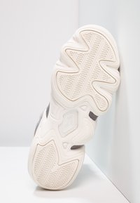 Podeszwa sneakersów o zakrzywionym, teksturowanym wzorze. Charakteryzuje się jasną kremową kolorystyką, z zygzakowym wzorem bieżnika i detalami brandingowymi.