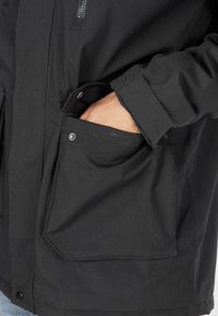 Svart jacka i ett texturerat material, med två stora framfickor, dragkedjestängning och knappar som dekoration på ärmarna.