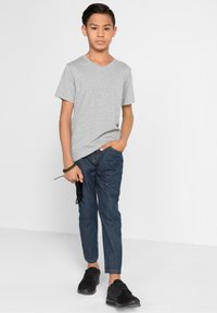 T-shirt grigia a collo a v abbinata a jeans blu scuro e scarpe sportive nere. Il modello tiene occhiali da sole in mano, in piedi davanti a uno sfondo bianco.
