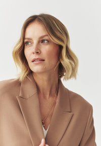 Blazer beige con spalle strutturate, una texture liscia e una leggera lucentezza, indossato sopra un top bianco con una delicata collana d'oro.