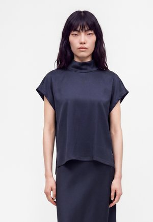 Femme aux cheveux raides et foncés portant un haut noir à manches courtes et col montant ainsi qu’une jupe assortie sur un fond clair uni.