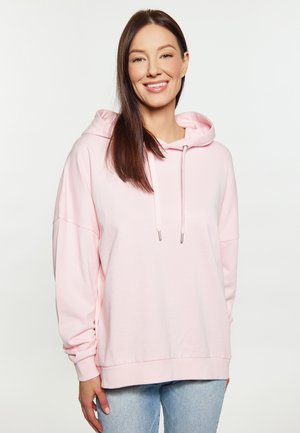 Lächelnde Frau mit langen braunen Haaren, die einen hellrosa Hoodie und hellblaue Jeans vor einem einfachen weißen Hintergrund trägt.