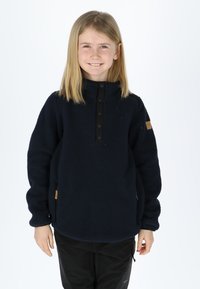Pullover in pile blu navy con colletto alto, chiusura con bottoni a pressione e tasche laterali. Texture morbida e design minimalista con un'etichetta a forma di patch marrone.