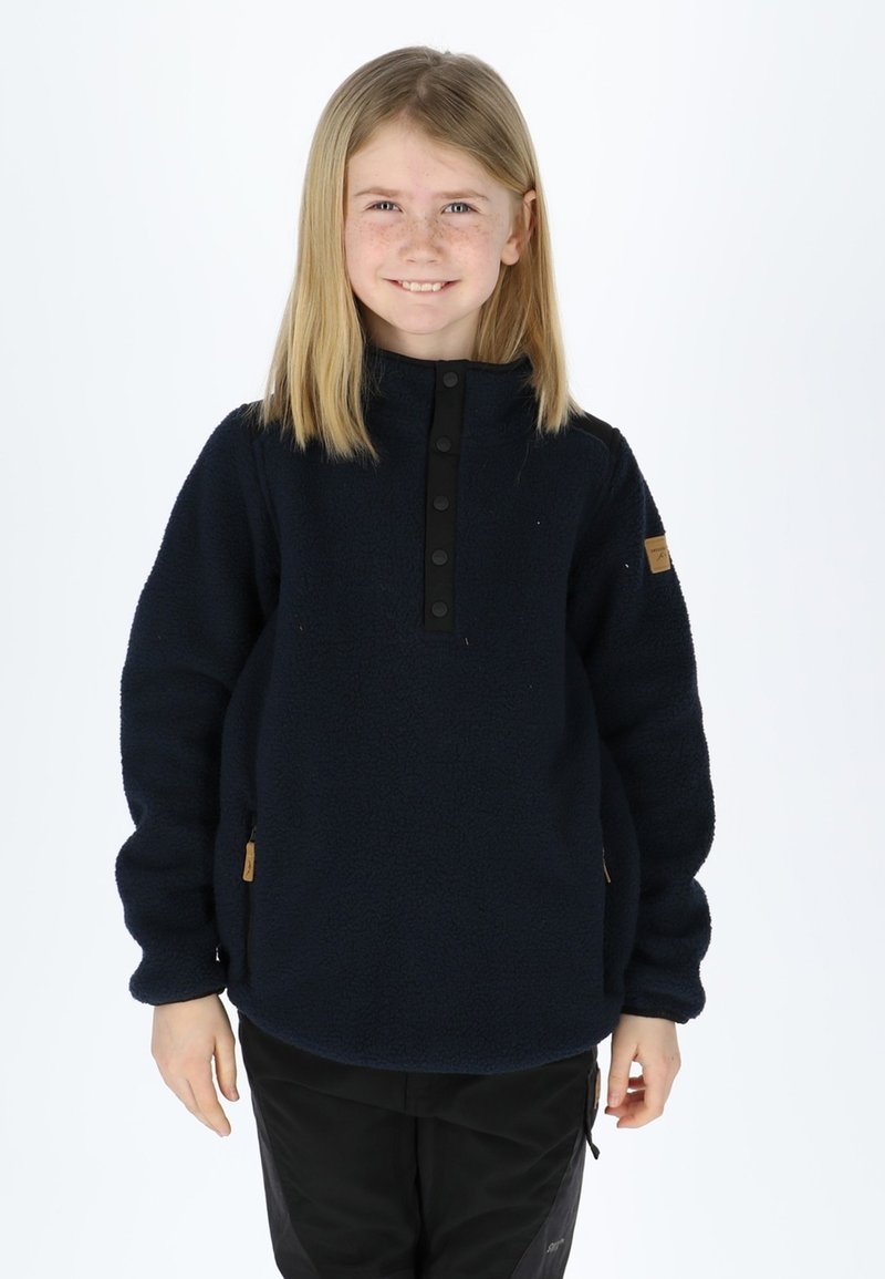 Pullover in pile blu navy con colletto alto, chiusura con bottoni a pressione e tasche laterali. Texture morbida e design minimalista con un'etichetta a forma di patch marrone.