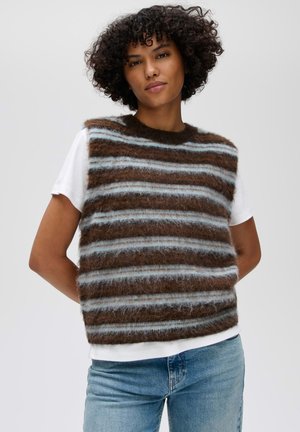 Vrouw met krullend donker haar draagt een zachte bruine en lichtblauwe gestreepte mouwloze trui over een wit T-shirt met korte mouwen en blauwe jeans.