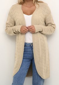 Femme portant un cardigan en tricot beige par-dessus un haut blanc et un jean bleu, debout devant un mur blanc uni.