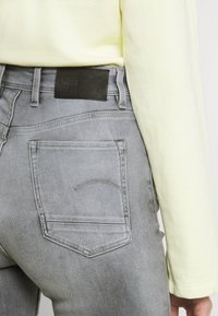 Jeans de mezclilla gris con una silueta ajustada, que cuentan con bolsillos traseros y un parche de cuero negro en la cintura. Camisa amarilla de manga larga visible.