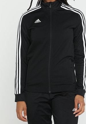 Persoon die een zwart Adidas trainingspak draagt met witte strepen op mouwen en broek, staand tegen een effen lichte achtergrond.