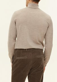 Beiger Langarmpullover mit hohem Kragen, aus weichem Stoff. Kombiniert mit dunkelbraunen Cordhosen mit Leistentaschen.