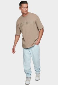 Camiseta oversized beige con texto gráfico, combinada con jeans holgados de color azul claro y zapatillas blancas. Tela suave, corte relajado y escote redondo.