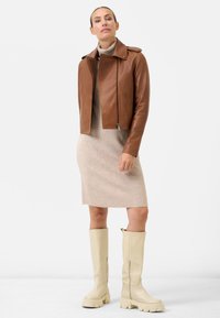 Bruin leren motorjack met een kraag, een beige gebreide jurk en crèmekleurige laarzen tot de knie met dikke zolen. Eenvoudig, modern ontwerp.