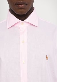 Světle růžová pruhovaná košile s rozšířeným límcem, která má na hrudi malé vyšívané logo a béžové knoflíky. Texturovaná tkanina.