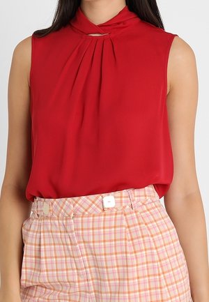 Blouse rouge sans manches avec un col plissé, associée à un pantalon taille haute à carreaux roses et oranges, orné de deux boutons blancs.
