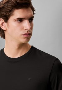 T-shirt preta de algodão com gola redonda e logótipo subtil cK. Apresenta mangas curtas e uma textura suave. Design simples, sem detalhes adicionais.