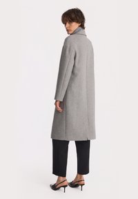 Kobieta z krótkimi kręconymi włosami, ubrana w szary długi płaszcz, czarne krótkie spodnie oraz czarne buty na obcasie z odkrytymi piętami, stoi i patrzy przez ramię.