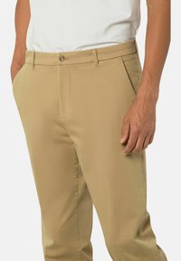 Beige Hose mit glatter Textur, ausgestattet mit einem Knopfverschluss und Seitentaschen, gezeigt an einem Model, das ein weißes Hemd trägt.