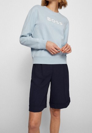 Personne portant un sweat-shirt bleu clair avec le texte "BOSS" et un short sombre jusqu'aux genoux, debout devant un fond uni.