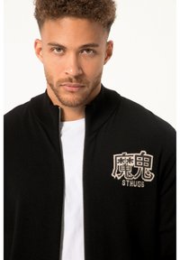 Sort zip-up sweater med ribbet krave, prydet med et elfenbensfarvet broderet logo på venstre bryst, båret over en hvid T-shirt.