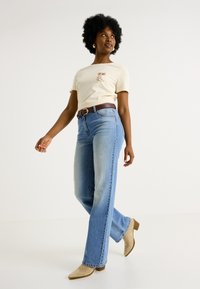 Camiseta beige con estampado gráfico, jeans de pierna ancha de color azul claro, cinturón marrón y botas de tobillo beige. Diseño sencillo, textura suave y estilo informal.