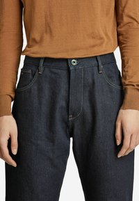 Jeans in denim blu scuro con vestibilità dritta, design a cinque tasche, cuciture visibili e chiusura a bottone verde. Abbinati a un top marrone a maniche lunghe.