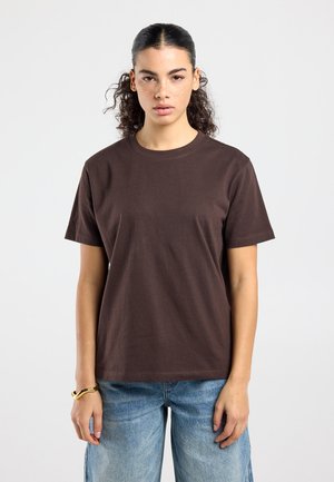 Jongere met krullend haar in een knot, gekleed in een eenvoudige donkerbruine korte mouwen shirt en lichtblauwe spijkerbroek, staand tegen een witte achtergrond.