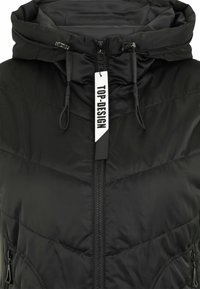 Schwarze gepolsterte Weste mit hohem Kragen und verstellbarem Kapuze. Verfügt über einen Reißverschluss vorne, seitliche Taschen und ein Etikett mit der Aufschrift "TOP-DESIGN."