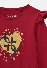 Camicia rossa a maniche lunghe realizzata in cotone biologico, con un design grafico oro e nero a forma di cuore con stelle e dettagli arricciati sulle spalle.