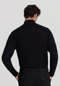 Pullover nero a maniche lunghe con collo alto, realizzato in materiale morbido. Presenta polsini a costine e una silhouette aderente, indossato con una cintura texturizzata.