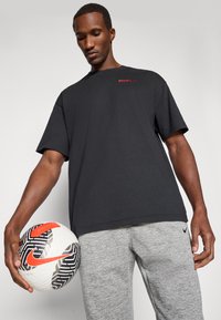 Czarny bawełniany t-shirt z czerwonym logo "ATLETI", zestawiony z szarymi spodniami sportowymi. Osoba trzyma wzorzystą piłkę do futbolu z pomarańczowymi akcentami.