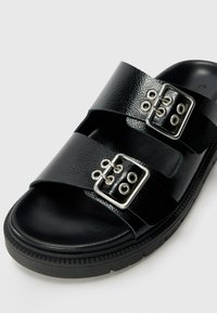 Sandales ouvertes noires avec deux larges lanières, ornées de boucles en argent et d'œillets, fabriquées en matériau texturé, avec une semelle en caoutchouc durable.