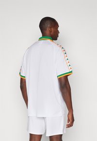 Polo-shirt de sport blanc avec des accents verts et orange, présentant des détails de logo sur les côtés, des manches courtes et une coupe slim, athlétique.
