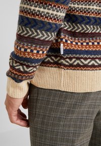 De gebreide trui heeft veelkleurige geometrische patronen in bruin, blauw en grijs, met een crèmekleurige ribbelrand en een zichtbaar merklabel.