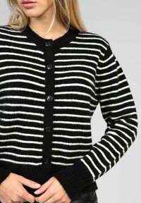 Cardigan rayé noir et blanc en tissu doux ; possède un col rond, des manches longues et cinq boutons noirs sur le devant.