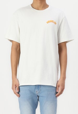 Mężczyzna ubrany w gładki, kremowo-biały t-shirt z krótkim rękawem, z małym pomarańczowym logo "WRANGLER" na lewej piersi i jasnoniebieskie dżinsy.