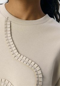 Sweat-shirt beige avec détail de ruban texturé sur le côté, encolure ronde et tissu lisse. Le design présente une forme unique et fluide.