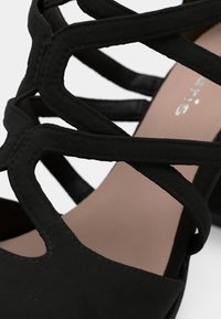 Μαύρο σανδάλι με λουράκια, κλειστό τακούνι και ταμπά πάτο, με μερικό ασημένιο κείμενο μάρκας ορατό.