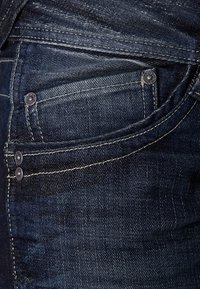 Mörkblå denimjeans med en framficka, synlig söm i en ljusare tråd och metallnitar för förstärkning.