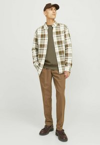 Férfi barna sapkában, olajzöld ingben, nyitott bézs és barna kockás dzsekiben, barna nadrágban és sötétbarna cipőben áll, és jobbra néz.