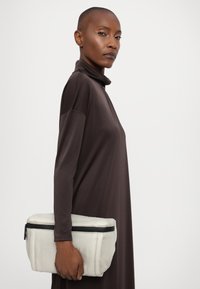 Clutch bianco texturizzato con chiusura a zip, tenuto in una mano, su uno sfondo semplice, indossato con un vestito marrone a collo alto.