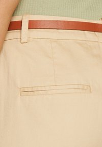 Beige broek met een gladde textuur, voorzien van een horizontale achterzak, aangevuld met een bruine leren riem.