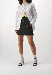 Chemise blanche boutonnée à l'avant, portée ouverte sur une jupe noire avec un cordon de serrage vert néon. Talons noirs à brides avec des accents vert vif.