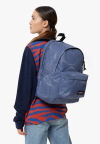 Zaino blu con texture, dotato di un compartimento principale e una tasca frontale, caratterizzato da un patch con logo e spallacci regolabili.