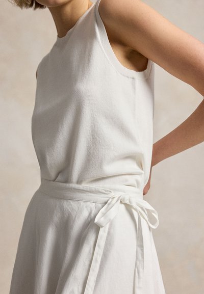 Moteris vilki baltą rankovių suknią su audinio diržu, surištu kaspinu ties liemeniu, stovinti su ranka ant klubo neutralaus fono.