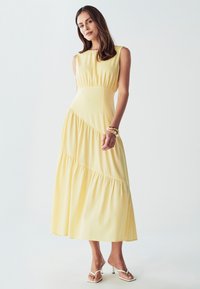 Rumena midi obleka s prilegajočim bodicem in večplastno, nabirano krilo. Narejena je iz mehke tkanine z gladko teksturo in brez rokavov.